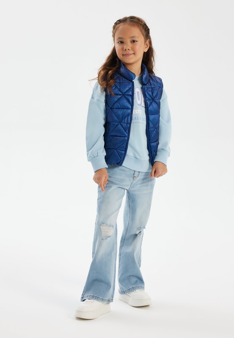 Blau gesteppte Weste über einem hellblauen Sweatshirt, kombiniert mit hellblauen Jeans im Schlag mit Rissen und weißen Sneakern.