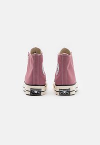 Růžové vysoké tenisky z plátna s bílým šitím, gumovou podrážkou a černým nápisem 'Converse All Star' na zadní straně. Klasický design na šněrování.