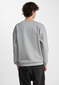 Sudadera gris de tela suave, con cuello redondo y puños acanalados, diseñada con un corte relajado y hombros caídos, usada con jeans oscuros.