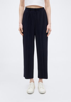 Ženska, ki nosi črne plisirane široke hlače, beli kratki top in bele superge, stoji na enostavni svetli podlagi.