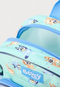 Ανοιχτό μπλε σακίδιο πλάτης με χαρακτήρες καρτούν σκύλων. Δύο θήκες με φερμουάρ, υφασμάτινη υφή με πολύχρωμο μοτίβο κομφετί. Εμφανίζεται το λογότυπο "BLUEY".