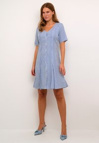 Vestito a maniche corte a righe blu e bianche con scollatura a V, bottoni frontali, orlo svasato e tessuto leggero. Abbinato a scarpe con tacco blu chiaro.