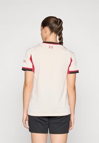 Kortærmet sportsjersey i lys beige med røde detaljer og sorte kanter. Ensfarvet ryg, fremstillet af åndbart materiale, parret med sorte shorts.