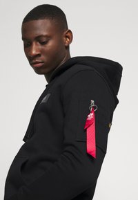 Black hoodie made of a soft material, featuring a zip pocket on the left arm with a red pull tab and a small logo on the chest.