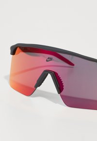 Nike-Brille mit einem dunklen Rahmen und einem Verlauf von Rot zu Orange bei den Gläsern. Sie verfügt über ein umschließendes Design mit einer strukturierten Nasenbrücke für besseren Halt.