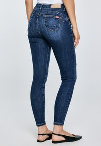 Asmuo dėvintis tamsiai mėlynas aptemptas džinsus ir juodus atvirus batelius, stovintis priešais šviesią, paprastą foną.