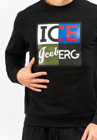 Crna dukserica s hrabrim, višebojnim grafičkim dizajnom na kojem se nalazi riječ "ICE" u crvenoj i plavoj boji, te "Iceberg" u bijelim i zelenim slovima.