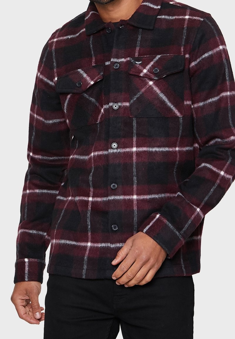 Homme portant une chemise à carreaux bordeaux, noire et blanche avec des poches poitrine, assortie à un pantalon noir, debout devant un fond uni.