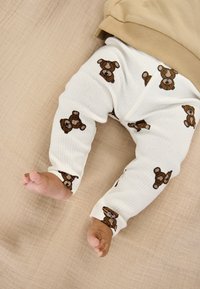 Hvide ribbede leggings med brune bamseprints, kombineret med en beige top. Synlige babyfødder, der hviler på en struktureret, neutral overflade.