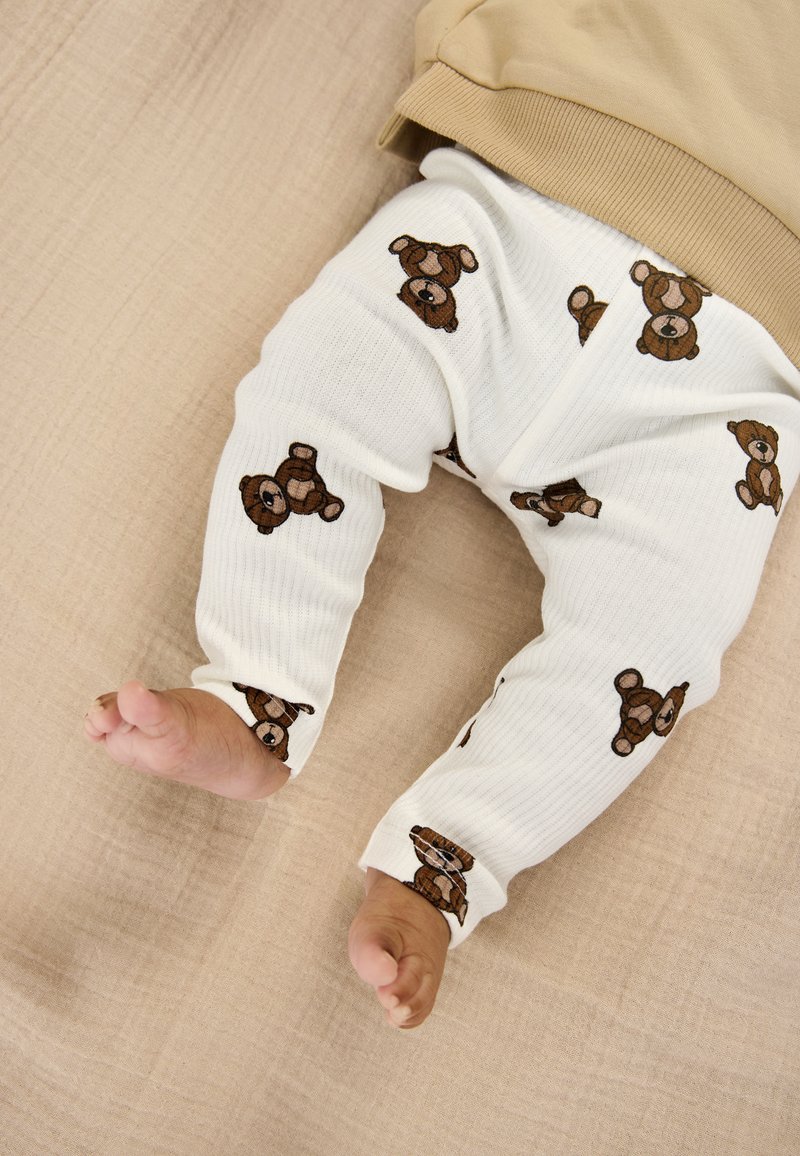 Hvide ribbede leggings med brune bamseprints, kombineret med en beige top. Synlige babyfødder, der hviler på en struktureret, neutral overflade.