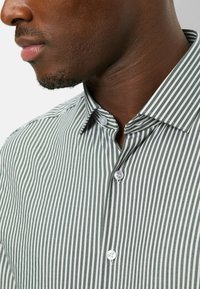 Grün-weiß gestreiftes Hemd mit Knöpfen, das einen Kragen, kurze Ärmel, glattes Gewebe und weiße Knöpfe an der vorderen Knopfleiste aufweist.