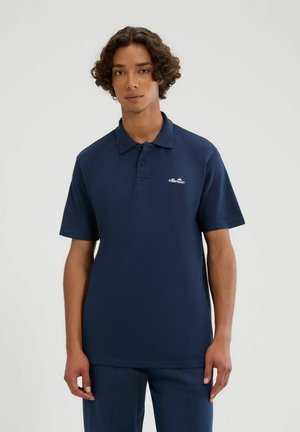 Młody mężczyzna z kręconymi włosami, ubrany w granatową koszulkę polo ellesse i pasujące spodnie, na jednolitym jasnym tle.