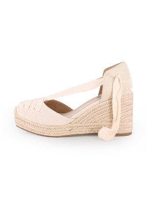 Cuña de esparto beige con parte superior de tela trenzada, tiras cruzadas en el tobillo y suela de yute texturizado. Presenta un detalle enrollado en el talón.