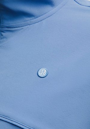 Jasnoniebieska tkanina z małym, okrągłym, haftowanym białym logo z stylizowaną literą „A” pośrodku.