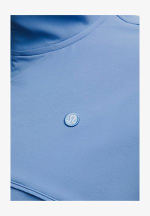 Jasnoniebieska tkanina z małym, okrągłym, haftowanym białym logo z stylizowaną literą „A” pośrodku.