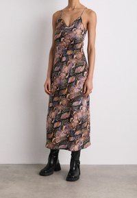 Femme portant une robe sans manches imprimée serpent, de longueur cheville, associée à des bottes à talons mi-mollet noires, se tenant contre un mur blanc.