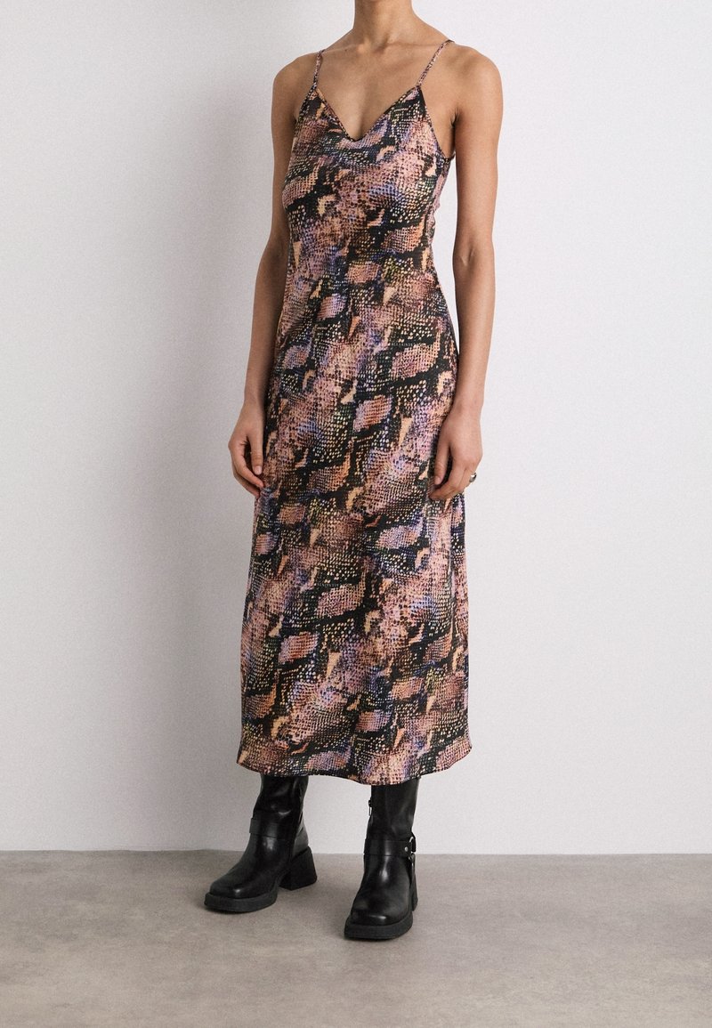 Femme portant une robe sans manches imprimée serpent, de longueur cheville, associée à des bottes à talons mi-mollet noires, se tenant contre un mur blanc.