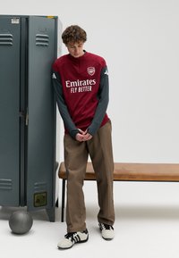 Maillot de sport rouge à manches longues grises, avec le logo "Emirates". Pantalon beige et chaussures de sport noires et blanches, debout à côté d'un casier.