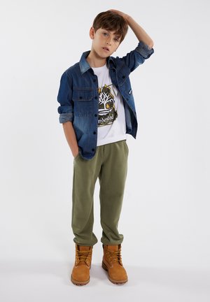 Spijkerjack, wit graphic t-shirt, olijfgroene sweaterbroek en tan veterschoenen. Casual outfit met verschillende texturen en kleuren.