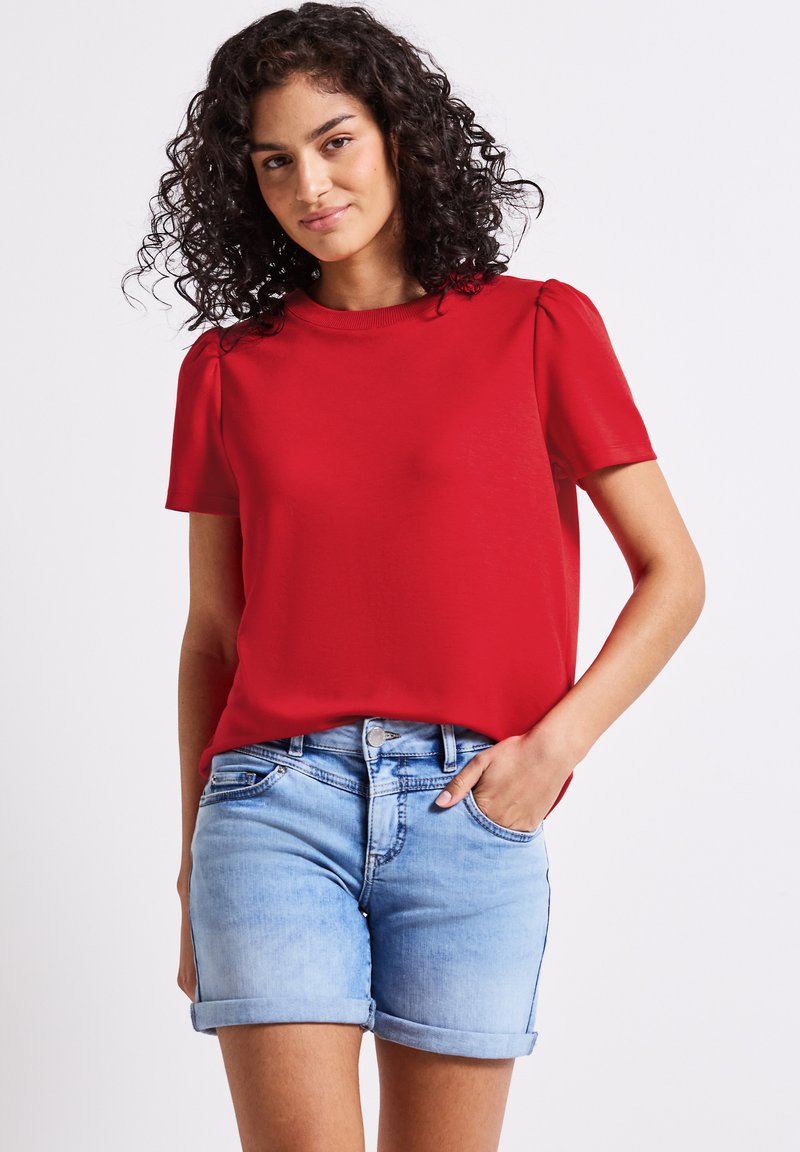 Top rosso a maniche corte con scollo rotondo e leggere maniche a sbuffo, abbinato a pantaloni shorts in denim azzurro chiaro con orlo arrotolato.