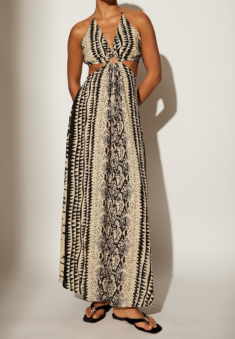 Femme portant une robe maxi sans manches à motifs, à col halter, avec des découpes et un détail anneau central, chaussée de sandales plates noires sur un fond uni.