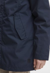 Navy waterproof parka with a textured fabric, featuring a side pocket and a square logo tag at the hem. Long sleeves with a cuff finish.