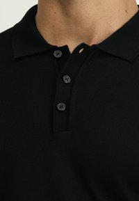 Schwarzes Poloshirt mit gerippter Textur, das drei schwarze Knöpfe am Kragen und ein Knopfloch aufweist.