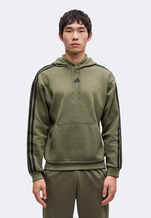 Jeune homme portant un sweat à capuche vert olive et un pantalon assorti avec des bandes noires le long des manches et des côtés, debout devant un fond uni.