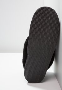 Chausson noir avec semelle texturée et intérieur moelleux appuyé contre un mur blanc sur une surface blanche.