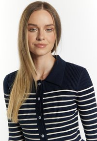 Gestreifter Cardigan in Marineblau und Weiß mit gerippter Struktur, ausgestattet mit einem Kragen und Vorderknöpfen, an einem Model vor einem weißen Hintergrund gezeigt.