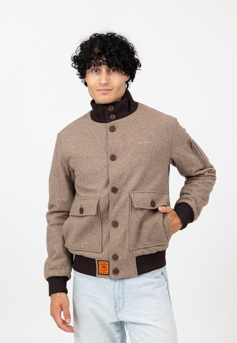 Mann mit lockigem schwarzen Haar, der eine braune Jacke mit Knöpfen und Taschen sowie einem orangefarbenen Etikett trägt, dazu hellblaue Jeans, steht mit einer Hand in der Tasche.
