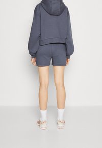 Sweat à capuche court gris foncé avec manches, assorti à un short coordonné. Le modèle porte des chaussettes blanches et des chaussures de sport de couleur claire.