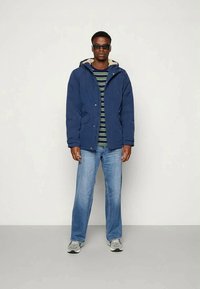 Tumši zila puffer jaka ar kapuci, apvienota ar svītrainu krekliņu. Gaiši zili džinsi un pelēki sporta apavi noslēdz apģērbu.