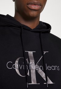 Felpa nera con cappuccio che presenta un grande logo argento "Calvin Klein Jeans" sul davanti, con cappuccio regolabile e colletto a costine.