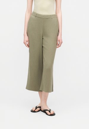 Donna che indossa pantaloni a gamba larga a vita alta di colore verde oliva, tagliati sopra la caviglia, abbinati a sandali infradito neri e una canottiera beige senza maniche.