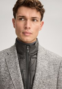 Grauer Blazer mit Muster über einem schwarzen Stehkragen mit Reißverschluss, mit glatter Textur und strukturierter Gestaltung. Dünne Revers und sichtbare Brusttasche.