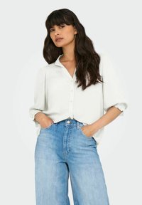 Hvid skjorte med knapper og opfoldede ærmer, parret med lyse denim jeans med høj talje. Stoffet ser glat og let ud.