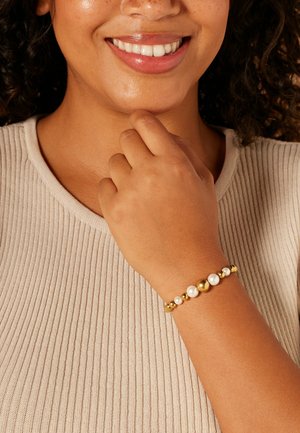 Gold and pearl bracelet featuring alternating gold and white spherical beads with varying sizes, set against a textured beige attire.