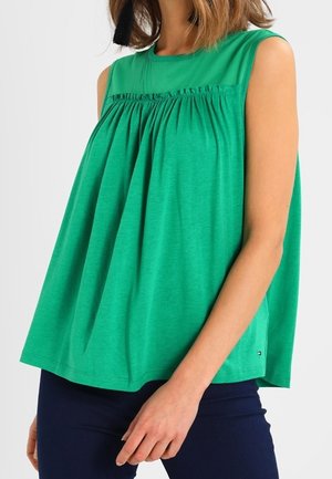 Mujer con una blusa verde sin mangas y suelta con pliegues sobre el pecho y pantalones azul marino, mano apoyada al lado.