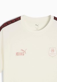 Tricou alb cu mânecă scurtă, având accente roșii cu un model pe umeri. Are logo-ul roz "Puma" și textul "KING", plus emblema "Stade Rennais".