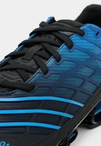 Črno in modro atletsko obuvalo z teksturirano zgornjo površino, ki ima valovite vzorce, črne vezalke in debel podplat z modrimi poudarki.