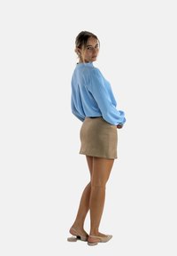 Blusa strutturata di un azzurro chiaro con maniche lunghe, abbinata a una mini gonna in suede beige. Modella che indossa scarpe beige senza chiusura, in piedi di lato.