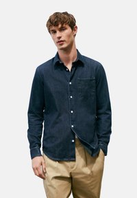 Donkerdenim overhemd met knopen en een borstzak, voorzien van witte knopen en een gebogen zoom. Gepaard met lichtgekleurde broek.