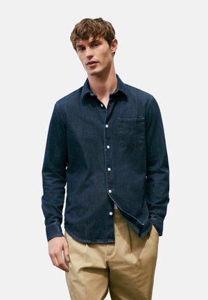 Donkerdenim overhemd met knopen en een borstzak, voorzien van witte knopen en een gebogen zoom. Gepaard met lichtgekleurde broek.