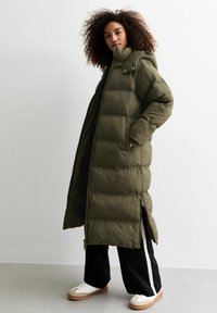 Lång, olivgrön puffjacka med dragkedja och huva, med en quiltad design och sidodragkedjor, tillsammans med svarta byxor och sneakers.