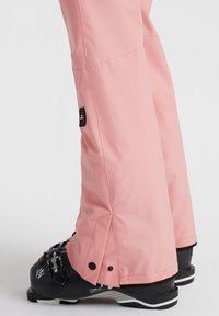 Pink ski pants with a smooth texture, featuring a fitted shape, adjustable bottom with snaps, and paired with black ski boots.