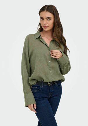 Nő laza, olíva zöld, gombos ingben és sötétkék farmerral, hosszú barna hajjal, enyhén megérintve az ing gallérját.