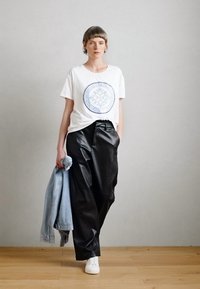 Λευκή μπλούζα με μπλε κυκλικό γραφικό, μαύρα δερμάτινα παντελόνια και ανοιχτό τζιν μπουφάν σε έναν ώμο; λευκά αθλητικά παπούτσια. Λείες υφές, χαλαρός σχεδιασμός.