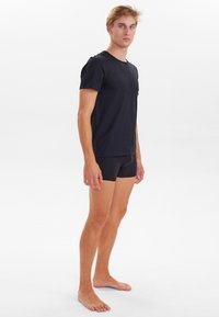 Sort bomulds T-shirt med korte ærmer, der står i kontrast til sorte tætsiddende shorts. Modellen står barfodet på en neutral baggrund.