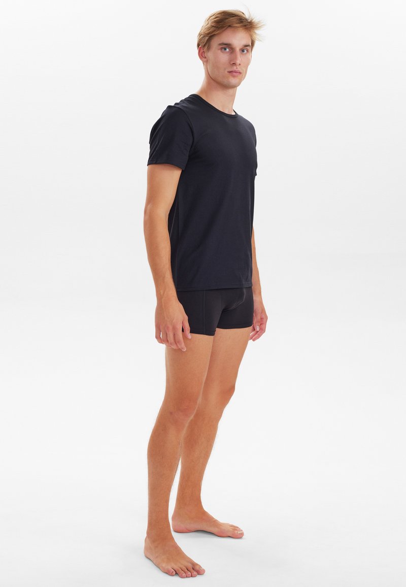 Sort bomulds T-shirt med korte ærmer, der står i kontrast til sorte tætsiddende shorts. Modellen står barfodet på en neutral baggrund.